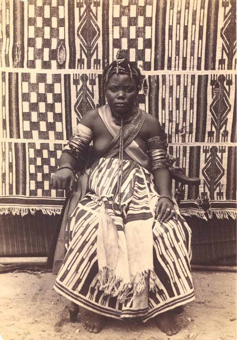 A black and white photographic print from c. 1880 by J.A. Green. The image depicts a young adult female seated in a chair, adorned in Igbo wedding attire. She wears a patterned textile dress, elaborate beaded jewelry, and a unique hairstyle. A patterned textile serves as the backdrop. (British Museum, Af,A50.3)