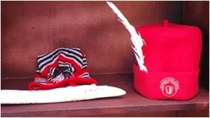 A bright red Igbo cap (Okpu Mme) with intricate beadwork and feathers, symbolizing spiritual and cultural authority, displayed on a wooden shelf beside a black and white patterned cap.